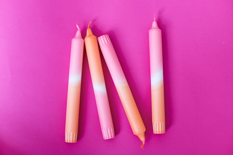 May include: Four ombre candles in shades of pink and orange. The candles are arranged in a row on a pink background.