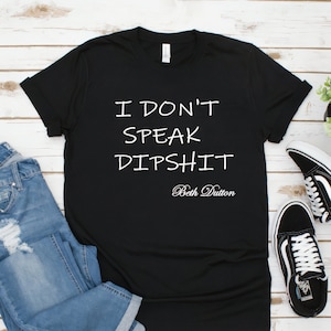 May include: Black t-shirt with white text that reads "I don't speak dipshit Beth Dutton". The shirt is on a white wood background with a pair of blue jeans and black sneakers.