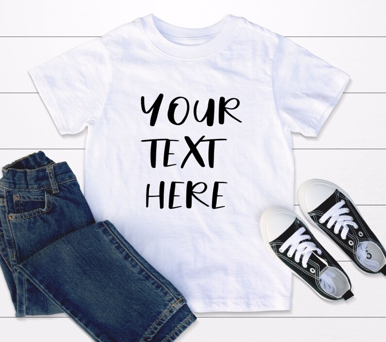 May include: White t-shirt with black text that says "YOUR TEXT HERE" on a white wooden background. The t-shirt is lying on top of a pair of blue jeans and black trainers.