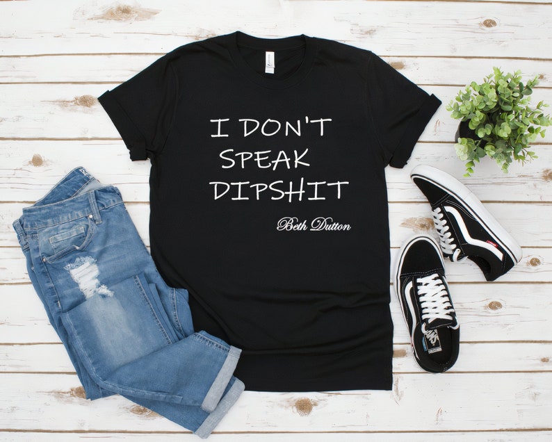 May include: Black t-shirt with white text that reads "I don't speak dipshit Beth Dutton". The shirt is on a white wood background with a pair of blue jeans and black sneakers.