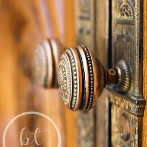 May include: Close-up of an antique door handle with a copper finish. The round doorknob features intricate detailing and a decorative design. The door and surrounding hardware are made of wood and metal.