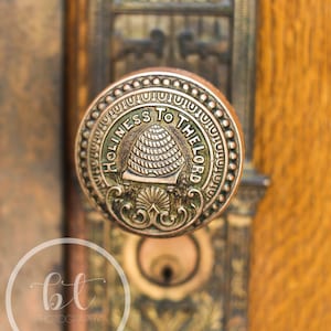 May include: A close-up of a brass door knob with a circular design. The knob features an inscription that reads "Holiness to the Lord" and a stylized beehive.
