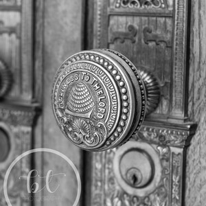 May include: Close-up of an ornate, antique door handle in black and white. The round handle features the words "HOLINESS TO THE LORD" around a beehive emblem. The door and surrounding frame are also intricately detailed.