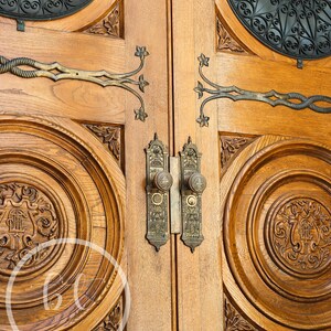 May include: Close-up of a pair of wooden doors with ornate carvings and brass hardware. The doors feature detailed circular designs, floral accents, and decorative handles. The wood has a warm, natural tone, and the metal hardware adds a touch of elegance.