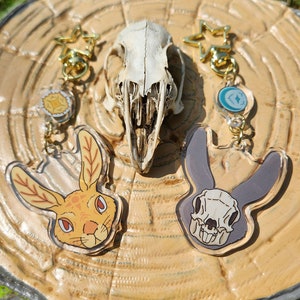 May include: Two acrylic keychains featuring a rabbit skull design. One keychain is a light brown rabbit skull with orange eyes and the other is a dark gray rabbit skull with white eyes. Both keychains are attached to gold star-shaped keyrings.