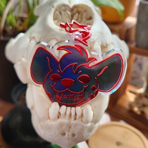 May include: A red and blue iridescent acrylic charm featuring a cartoon hyena with a toothy grin. The charm is attached to a silver ring.