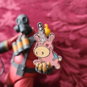 May include: A pink unicorn enamel pin with a silver outline. The unicorn has a white horn, brown hooves, and a yellow bird on its back. The pin is set against a red background.