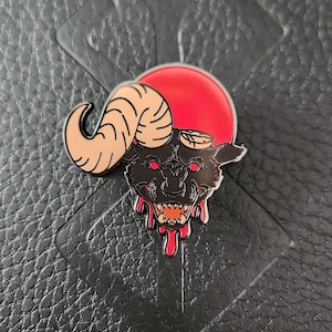 May include: A black enamel pin featuring a stylized wolf with ram horns, a red sun, and dripping blood details. The pin is set against a textured black surface.