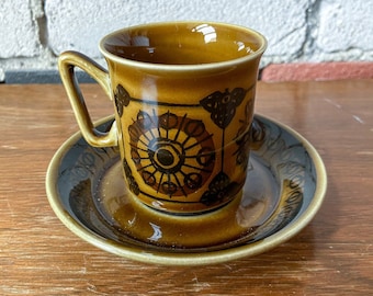 Puede incluir: Un juego de taza y platillo de cerámica marrón y negro con un patrón geométrico. La taza tiene un asa y el platillo tiene un borde festoneado.