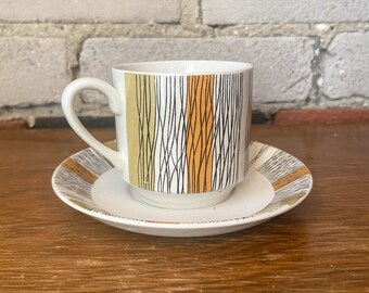 Puede incluir: Una taza de té y un platillo blancos con un patrón geométrico negro, blanco y amarillo. La taza y el platillo están sobre una superficie de madera.