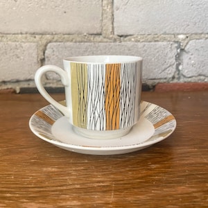 Könnte beinhalten: Eine weiße Teetasse und Untertasse mit einem geometrischen Muster in Schwarz, Weiß und Gelb. Tasse und Untertasse stehen auf einer Holzoberfläche.