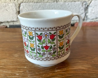 Puede incluir: Taza de cerámica blanca con un patrón floral en tonos rojos, amarillos, verdes y morados. El patrón presenta flores y hojas estilizadas dentro de un borde marrón.