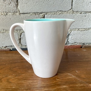 Puede incluir: Una jarra de cerámica blanca con un borde azul claro y un asa. La jarra está sobre una superficie de madera.