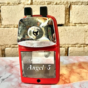 Puede incluir: Un sacapuntas rojo con la parte superior plateada y asas negras. El texto "Angel-5" se muestra en la parte delantera. El sacapuntas está sobre una superficie de mármol frente a una pared de ladrillos.