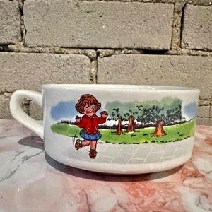 以下が含まれることがあります： 取っ手付きの白いセラミック製のスープボウル。ボウルには、赤いジャケットと青いスカートを着た女の子が木のある公園を歩いているという漫画のイラストが描かれています。