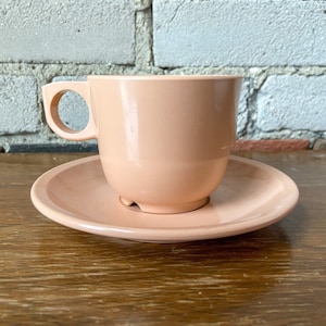 Puede incluir: Un juego de taza y platillo de color rosa p&aacute;lido. La taza tiene un asa redondeada y el platillo es ligeramente curvo.