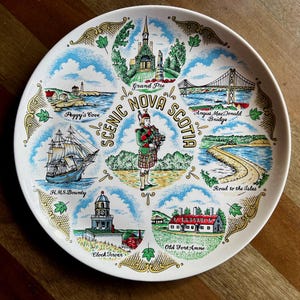 May include: A white decorative plate with illustrations of Nova Scotia landmarks, including Peggy's Cove and a bridge. The plate has a gold trim and the words "Scenic Nova Scotia". The plate features illustrations of a ship and other landmarks.