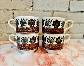 Puede incluir: Cuatro tazas de cerámica blanca con un diseño floral. Las tazas presentan un patrón repetido de flores y hojas verdes, marrones y rojas. Las tazas están apiladas en pares, con un asa blanca en cada taza.