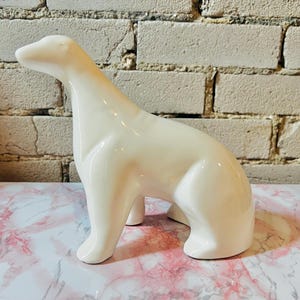 May include: A glossy white ceramic polar bear ornament. The bear is in a seated position, with its head raised and turned slightly to the left. The ornament has smooth, rounded features and is set against a brick wall.