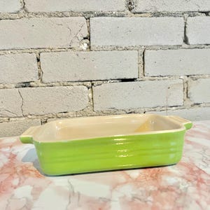 May include: A rectangular, light green ceramic baking dish with a cream-colored interior. The dish has a ribbed exterior and small handles on each end. The background features a white brick wall and a marble countertop.