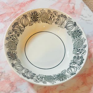 May include: White ceramic bowl featuring a black floral pattern around the rim. The bowl has a simple design with a black line around the inner circle. The floral pattern includes various flower and leaf shapes.