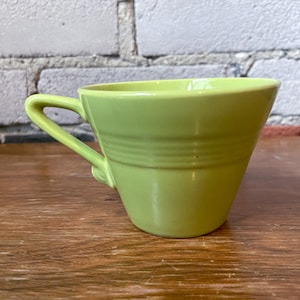 Puede incluir: Una taza de té de cerámica de color verde claro con forma redondeada y asa. La taza está sobre una superficie de madera.