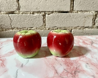 May include: Two ceramic apple-shaped salt and pepper shakers. The shakers are red with green tops and a glossy finish. They are sitting on a pink and white marble surface, with a brick wall in the background.
