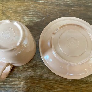 Puede incluir: Un juego de dos tazas de t&eacute; y platillos de cer&aacute;mica de color rosa claro. La parte inferior del platillo y la parte inferior de la taza de t&eacute; tienen ambos un logotipo circular que dice "Hecho en Canad&aacute;".
