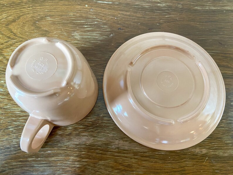 Puede incluir: Un juego de dos tazas de t&eacute; y platillos de cer&aacute;mica de color rosa claro. La parte inferior del platillo y la parte inferior de la taza de t&eacute; tienen ambos un logotipo circular que dice "Hecho en Canad&aacute;".