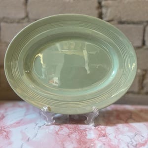 May include: A light green oval serving platter with a ridged rim. The platter is displayed on a clear plastic stand against a brick wall and a pink and white marble surface.