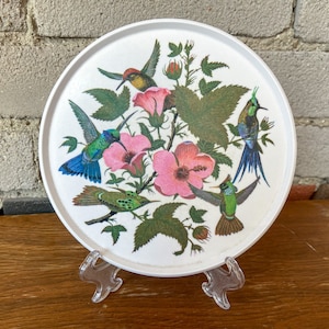 May include: A white, circular decorative tray with a vibrant illustration of hummingbirds and pink flowers. The tray showcases a detailed design with green leaves and various hummingbird species. The tray is displayed on a clear plastic stand.