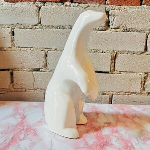 May include: A glossy white ceramic polar bear figurine. The sculpture has a smooth, curved design with the bear standing upright. The background features a brick wall and a pink and white marble surface.