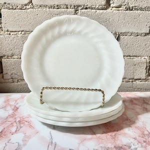 Puede incluir: Una pila de cuatro platos blancos de estilo vintage con un diseño de remolino. Los platos se exhiben en un soporte de metal dorado. Los platos están sobre una superficie de mármol rosa y blanco, con una pared de ladrillo blanco en el fondo.