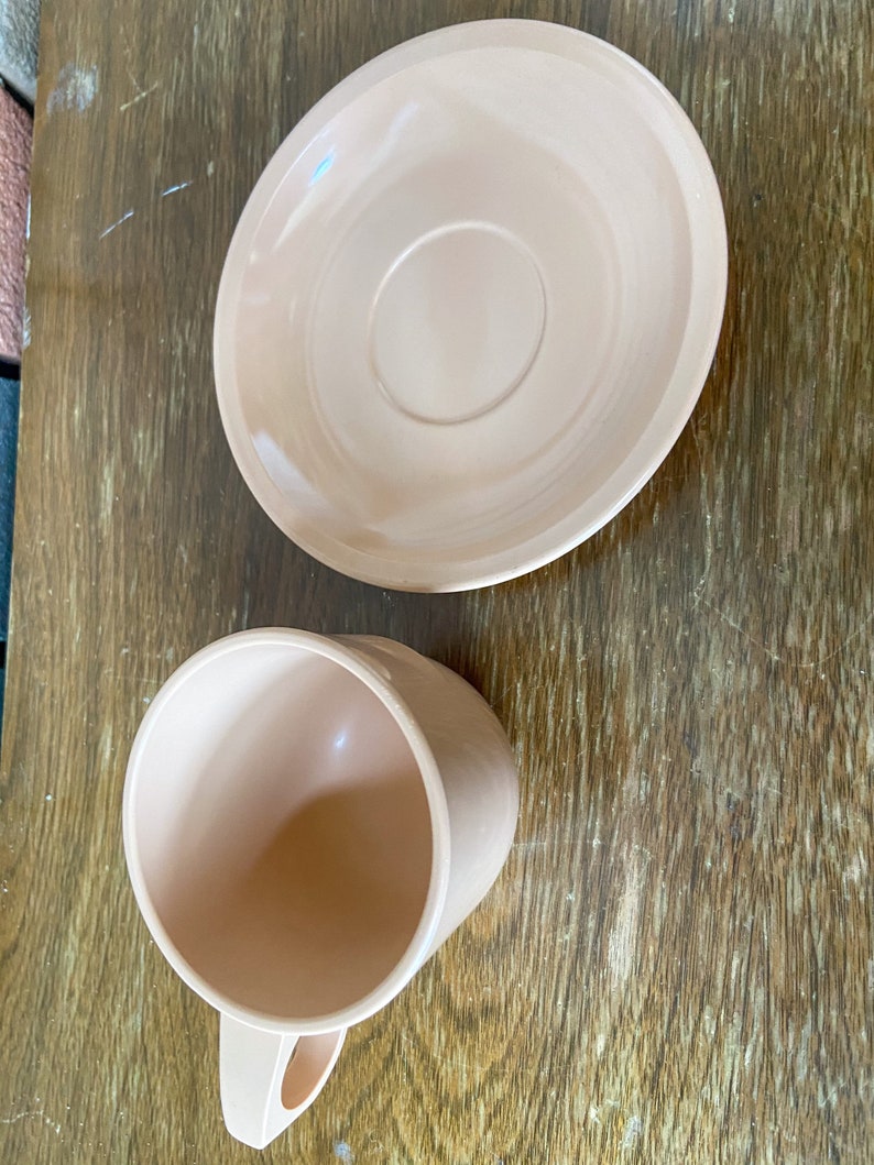 Puede incluir: Un juego de taza y platillo de color rosa p&aacute;lido. La taza tiene un asa redondeada y el platillo tiene un borde ligeramente elevado.