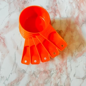 Puede incluir: Un conjunto de cinco tazas medidoras de color naranja brillante. Las tazas son de plástico y tienen mangos largos con marcas de medición. Las tazas están dispuestas en forma de abanico, con la taza más grande en la parte superior.