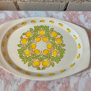 May include: Cream-colored oval serving platter with a speckled surface. The platter features a central design of yellow oranges and green leaves, surrounded by smaller orange and leaf motifs. A vintage-style serving dish.