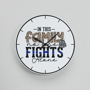 May include: White round clock with black hands and black text that reads "In this family no one fights alone". The clock has a blue and black thin line design around the edge and a leopard print design in the center. The clock is hanging on a white wall.