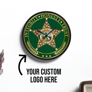 May include: A green and gold clock with a sheriff's star and the words "Integrity, Honesty, Trust" around the edge. The clock is on a white wall with a wooden shelf below it.