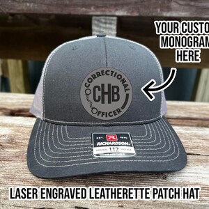 May include: A black and gray trucker hat with a gray mesh back. The hat has a black leatherette patch with the text "Correctional CHB Officer" and a chain link design. The hat is a Richardson 112 adjustable hat.
