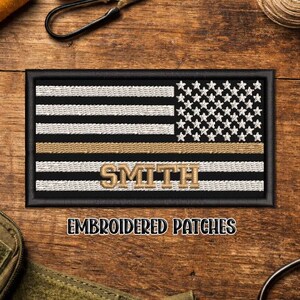 May include: Embroidered patch featuring a stylized American flag design with black and white stripes, a field of white stars, and a gold stripe. The name "SMITH" is embroidered in gold. The patch is framed in black.