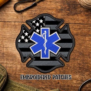 May include: Embroidered patch featuring a blue Star of Life emblem, a black and gray striped section, and a section with white stars on a black background. The patch is in the shape of a Maltese cross.