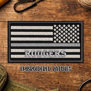 May include: Embroidered patch featuring a black and white American flag design with a gray stripe and the name "RODGERS" in gray letters. The patch is rectangular with a black border. The text "EMBROIDERED PATCHES" is below the patch.