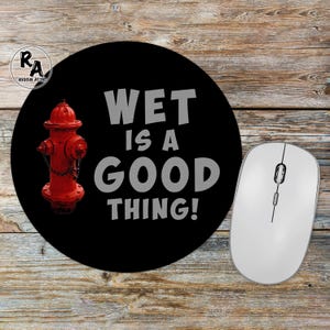 May include: Black round mousepad with the text "WET IS A GOOD THING!" and a red fire hydrant graphic. A white wireless mouse is to the right of the mousepad. The mousepad has a Rescue Attic logo in the upper left corner.
