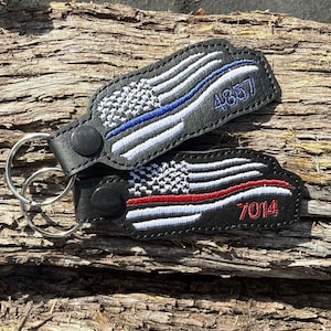 May include: Two black leather keyrings with embroidered American flags. One has a blue line and the number 4857, the other has a red line and the number 7014. Both have silver key rings and black snap closures.