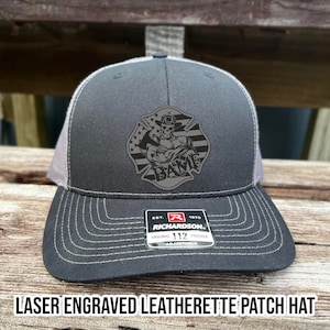 May include: A black and gray trucker hat with a laser engraved leatherette patch featuring a firefighter holding an axe. The patch is in the shape of a fire hydrant and has the text "EST. 1970 RICHARDSON 112 ADJUSTABLE".