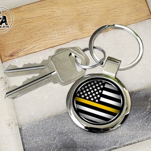 May include: A round silver keychain with a graphic of the American flag in black and white with a yellow stripe. Two silver keys are next to the keychain. The keychain is on a light gray and wood-grain surface.