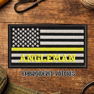 May include: Embroidered patch featuring a stylized American flag design with a yellow stripe and the word "ANGLEMAN" in yellow. The patch is rectangular with a black border. The text "EMBROIDERED PATCHES" is below.