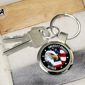 May include: A silver keychain with a round metal charm featuring a bald eagle with an American flag in the background. The charm has the text "America's Bravest" and "Retired" on it.