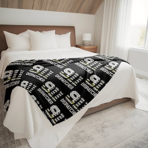 May include: Black and white throw blanket with a repeating pattern of the word "DISPATCHER" and a thin yellow stripe. The blanket is on a white bed with white pillows.