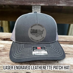 May include: A black and gray trucker hat with a laser engraved leatherette patch that reads "America's Bravest Correctional Officer Corrections Officer". The patch features a gray outline of the United States with an American flag design inside. The hat is adjustable and has a Richardson logo on the back.